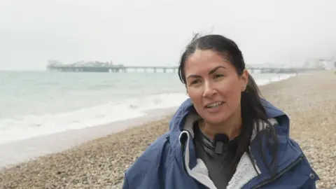 Peter Whittlesea / BBC A woman stood on a pebbly beach wearing a dry robe. A pier is visible behind her, jutting out from the water on the left hand side.