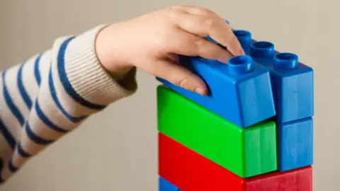 A child's hand stacking large building blocks. The blocks are blue green and red.