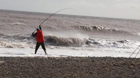 A man can be seen on a stony beach holding a fishing rod above the surf of a grey-brown sea near the shoreline. He has his back to the camera and is wearing a red jacket.