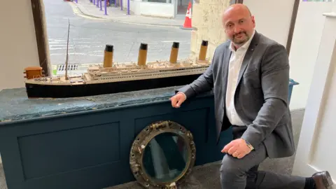 Andrew Aldridge is wearing a grey suit and white shirt and kneeling beside a replica model of the Titanic