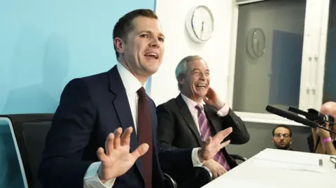 Robert Jenrick sits next to Nigel Farage at a desk in front of microphones. Both men are laughing. 