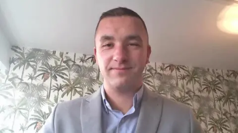 A screengrab from a Teams call, showing a man smiling at the camera standing in a room with wallpaper behind him.