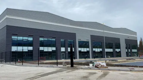 BAE Systems A grey warehouse with large windows along the front. The site looks empty, with a few items used in construction scattered around.