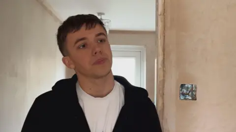 Nick Tait, standing in the corridor of his house, with bare plaster on the walls and what looks like an unfinished light socket on the wall. He has short hair and is wearing a white T-shirt and a black hoodie.