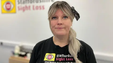 A woman with long blonde hair and a black bandana on her head, and wearing a black Staffordshire Sight Loss shirt, smiles as she stands in front of a white wall.