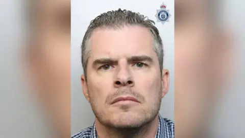 Mugshot of Simon Clark, 46, who has grey short hair and a grey beard. He frowns at the camera. 