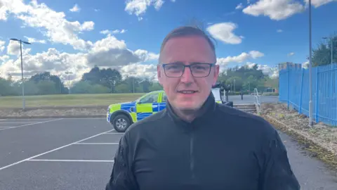 Photograph of PC Jonathan Markham-Barwell from Greater Manchester Police. He is pictured in a car park, with a police car behind him. 