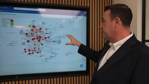 BBC/Al Candelin A man, Jonny, looks at a screen in a meeting room. On the screen are a number of coloured dots and blobs, linked by lines.