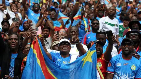 A crowd of DR Congo fans are seen in the stands cheering for their team during a 2026 Fifa World Cup play-off match against Jamaica. A fan in a white cap and blue shirt is in the foreground holding up a DR Congo flag, with many behind him holding up phones to take pictures