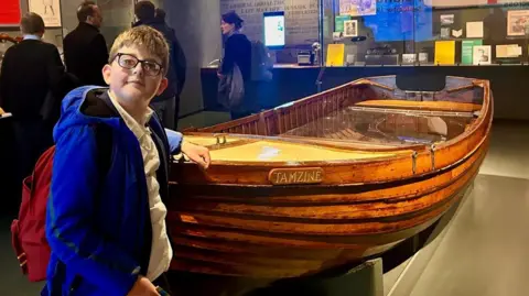 Family handout Ben, standing by a wooden boat, in a museum, wearing a blue jacket and red bag, on his back. Other people are in the room looking at displays. 
