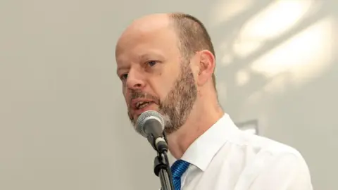 Hazel Plater Jamie Driscoll, who is wearing a white shirt and a blue tie, is speaking into a microphone. He is balding with very short cropped brown hair at the sides and a beard.