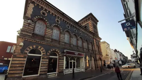 The orange and black building stands prominently on a street and a Costa coffee house can be seen inside the building. The upper level is boarded up and a person is walking down the street alongside the building.
