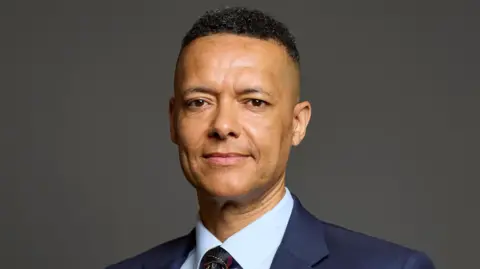 Clive Lewis looking at the camera wearing a blue jacket, a light blue shirt and a dark blue tie. He's looking at the camera with a slight smile on his face