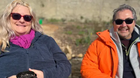 A women and a man are looking directly at the camera and smiling. The women is on the left and has blonde wavy hair, she is wearing a bright pink scarf and red sunglasses. She has a hot chocolate in her hand. The man is wearing a bright orange jacket over a grey zip up hoodie. He has black sunglasses and grey hair. 