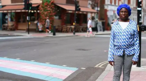 A woman stands on the side of a road in central London beside a road crossing painted in white, blue and pink stripes. 