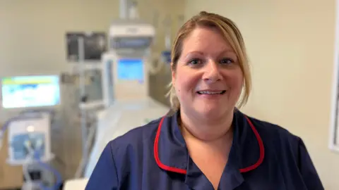 Francesca has blond hair, tied back, and wears a navy blue nurse's uniform. She is standing in front of an incubator.