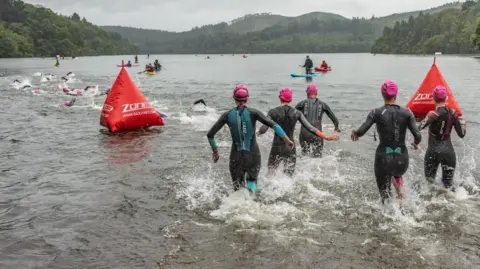 Five people in swimsuits are walking in a lake. They are all wearing pink swimming hats, and the water is up to just below their knees. Further away, several people are swimming. There are also a number of people on paddleboards. 