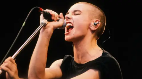 Getty Images Sinead O'Connor performing on stage, she has a shaved head and is wearing a black tshirt, she is gripping a microphone on a mic stand and singing passionately with her eyes closed.