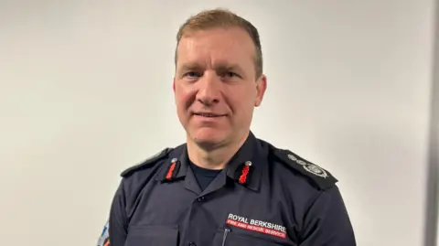 Wayne Bowcock is standing up wearing his RBFRS uniform, a blue shirt which has "Royal Berkshire Fire and Rescue Service" sewn into it.