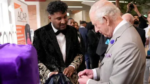 Getty Images Imran Khan stands talking to King Charles III. Mr Khan, on the left is wearing a black jacket, with gold embroidery on the sleeves, a white shirt and black bow tie. He is holding up the inside of another jacket up to show to the King, who is wearing a light grey suit and white shirt. Behind the two men a number of people are stood watching.