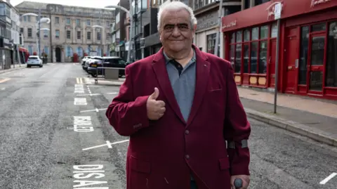 Campaigner Brian Roberts, with grey hair and wearing a maroon jacket, smiles and gives the "thumbs up" sign while standing next to new disabled parking bays