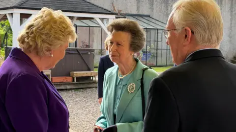 Princess Anne wearing a teal outfit. She is facing a woman who is wearing a purple top. There is also a man with white hair and glasses wearing a suit. 