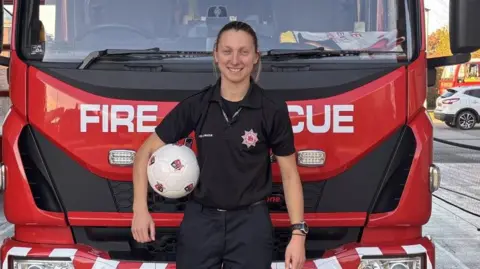 BBC A woman, Bow Jackson, wearing black firefighters' uniform, with a football under her right arm, standing in front of a red fire engine