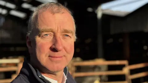 A close-up of Chair of the Royal Association of British Dairy Farmers, Robert Craig. He has short thinning white hair and bushy eyebrows. He is clean shaven. In the background is a cattle milking shed in soft focus.