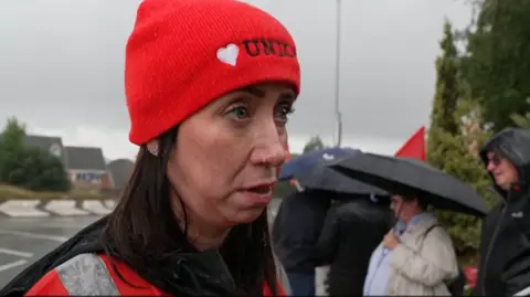 A woman in an orange hi-vis jacket and a red beanie hat with a heart and union written on it is being interviewed in the rain on a picket line. Several people holding umbrella and wearing raincoats can be seen blurred out on the picket line behind her.