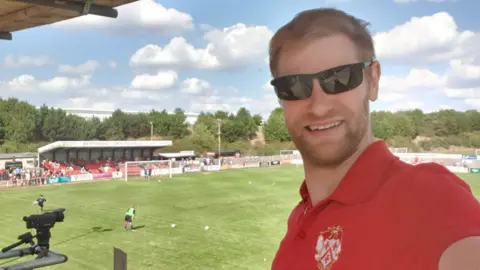Peter Short/@NorthantsLegend Peter Short standing on a gantry at Latimer Park. He is wearing black sunglasses and a red Kettering Town polo shirt with the club's crest on it. Behind him, the pitch is visible and players are warming up as people watch on from a small stand behind a goal.
