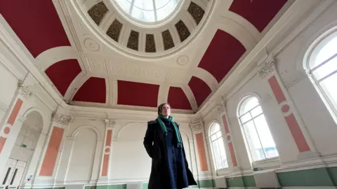 BBC A woman with short dark hair is wearing a navy blue winter coat and green scarf. She is stood in a derelict building which has an ornate ceiling with a glass dome and red paint. The walls are decorated with plaster and peach coloured paint