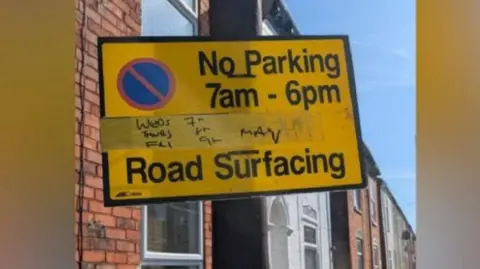 BBC A yellow sign attached to a lamppost with the words "No parking 7am to 6pm road surfacing"