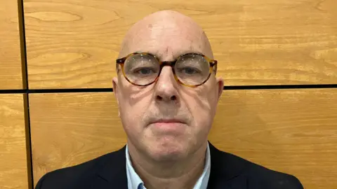 Stephen Maguire is looking straight into the camera. He is bald and has brown rimmed glasses. He is wearing a dark jacket and light blue shirt. There is a wooden wall behind him