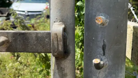 The image shows a broken lock on a metal fence. A car is partly visible in the background.