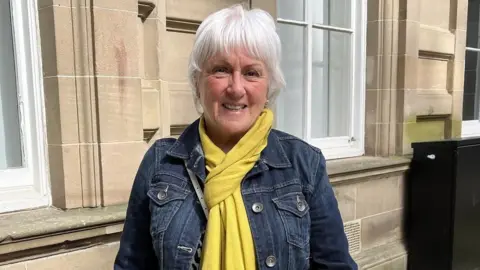 Marion Short is in her sixties with grey hair and wearing a denim jacket with a yellow scarf. She is standing in front of a stone building with two white windows 