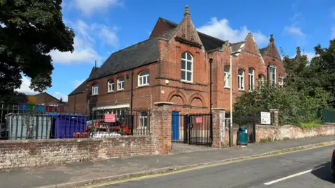 BBC A brick wall around Avenue Junior School, Norwich, with a gate, a sign saying Avenue Junior School to the right of the gate, and beyond the gate are large rubbish bins.