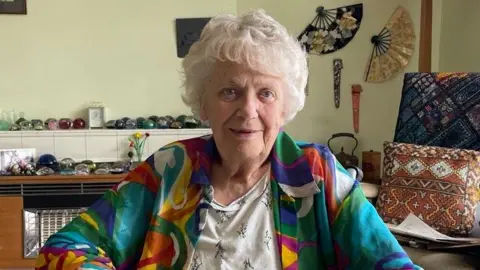 Sofia Luis-Hobbs/BBC An older person with short white hair sits indoors with hands resting together, wearing a colourful patterned jacket over a light top. The room contains shelves displaying decorative stones and ornaments, a heater, patterned cushions and wall-mounted decorative fans, creating a homely setting.