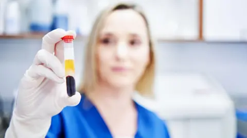 An in-focus hand in a white safety glove holds a vial between her thumb and index finger. It contains a red fluid, clearly blood and has a light red plastic lid. Out of focus in the background is a woman in a blue shirt.