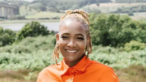 Vivian Tanaka smiles at the camera. She is on a hill that overlooks the River Orwell and Orwell Bridge near Ipswich. She wears a bright orange shirt and has blonde hair that has been braided and tied back.