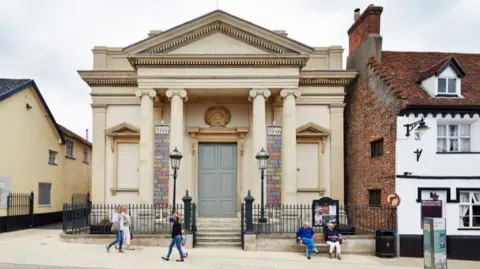 Getty Images A frontal view of Diss Corn Hall, which is a grand, neo-classical building, cream in colour. There are two old-fashioned lamps outside and black metal railings. Several people are walking past and a man and a woman are sitting on a bench outside.