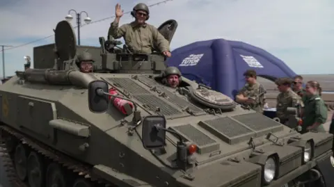 BBC A green military tank is driving down a street. Two men wearing helmet and sunglasses are peering through an open hatch at the top of the tank, while another is doing the same just in front. Other members in green military uniforms are standing by the side looking as it drives past.