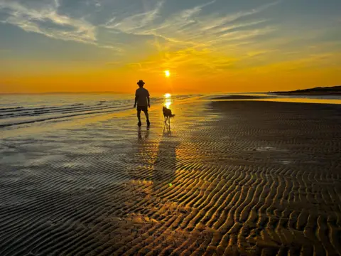 Jacki Gordon A silhouette of a man wearing a hat accompanied by a collie on a beach at sunset