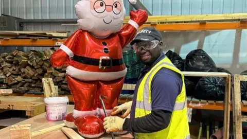 John Devine/BBC Saido is wearing a black baseball hat with the word 'Security' on it. He has a crowbar in his hand and is standing at a work bench, next to an inflatable Father Christmas. There are shelves behind him with strips of wood and filled-up bin bags. 