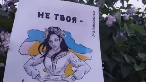 Um pôster mostra uma mulher claramente ucraniana em vestido tradicional e as mãos nos quadris olhando em desafio