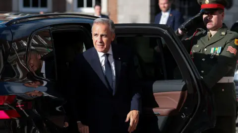 Reuters Carney steps out of a shiny black car. He wears a sharp suit. Behind him a solider salutes. 