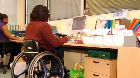 Man with dreadlocks sits at an office desk, in a blue wheelchair, typing at a desktop computer