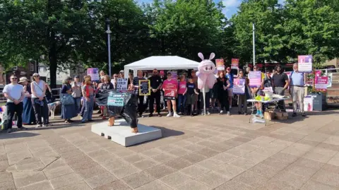 A sunny outdoor rally of people holding pink protest signs under a white canopy tent in a paved area. A pink mascot stands in the crowd, a small statue wrapped in black plastic sits in the foreground, and green trees line the background.