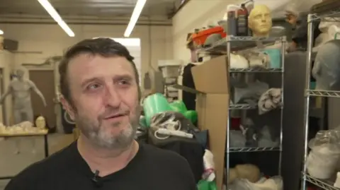 Mike Hill, wearing a black T-shirt, speaks to the BBC in his LA studio surrounded by masks on shelves.