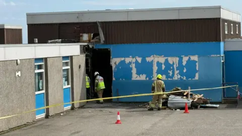 BBC Firefighters behind a yellow cordon investigate the damage to Dales Park primary school