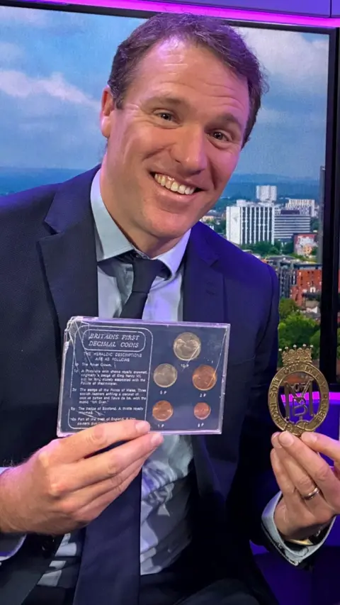 Ian Payne from Churchill Living holds up golden cypher of Lord Mountbatten and a display case with coins inside it. He sits in the Radio Solent studio wearing a dark blue suit, with a light blue shirt and dark blue tie.